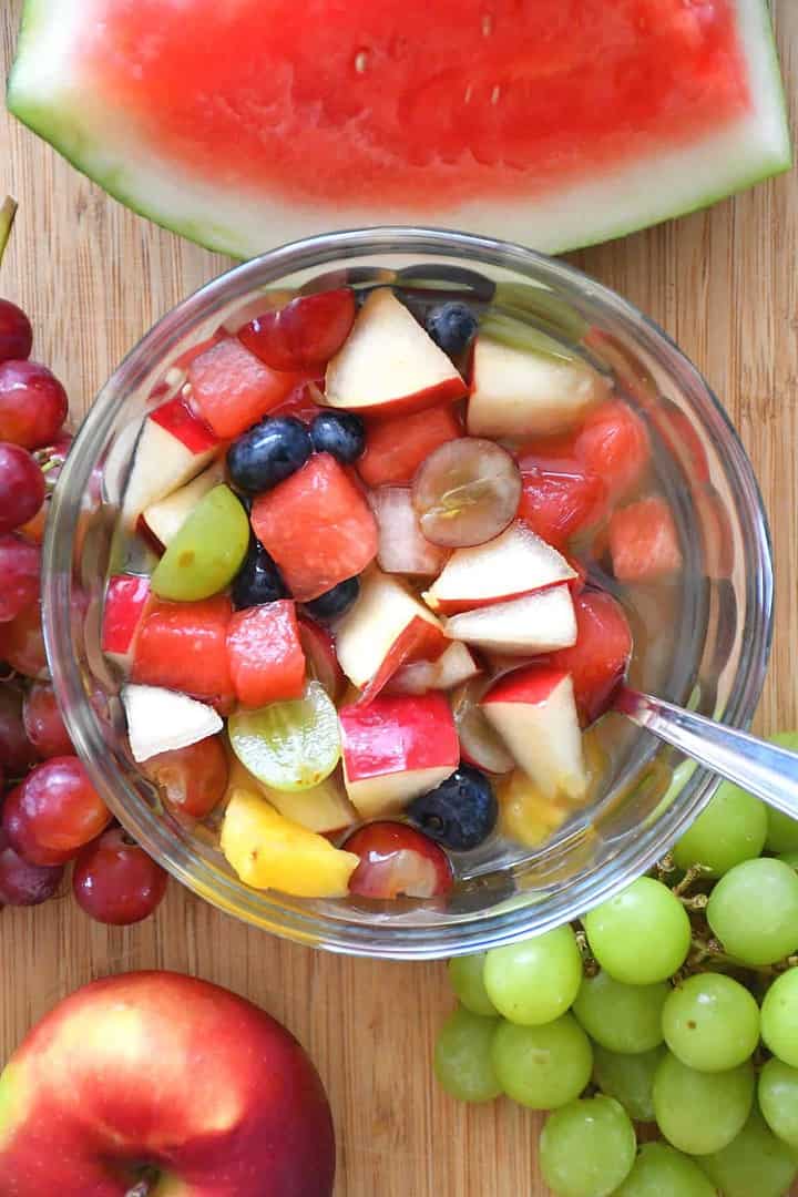 A healthy fruit salad featured on a bamboo board with red grapes, green grapes, an apple and a watermelon quarter.