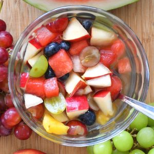 A kidney friendly healthy fruit salad featured on a bamboo board with grapes, watermelon and a apple.