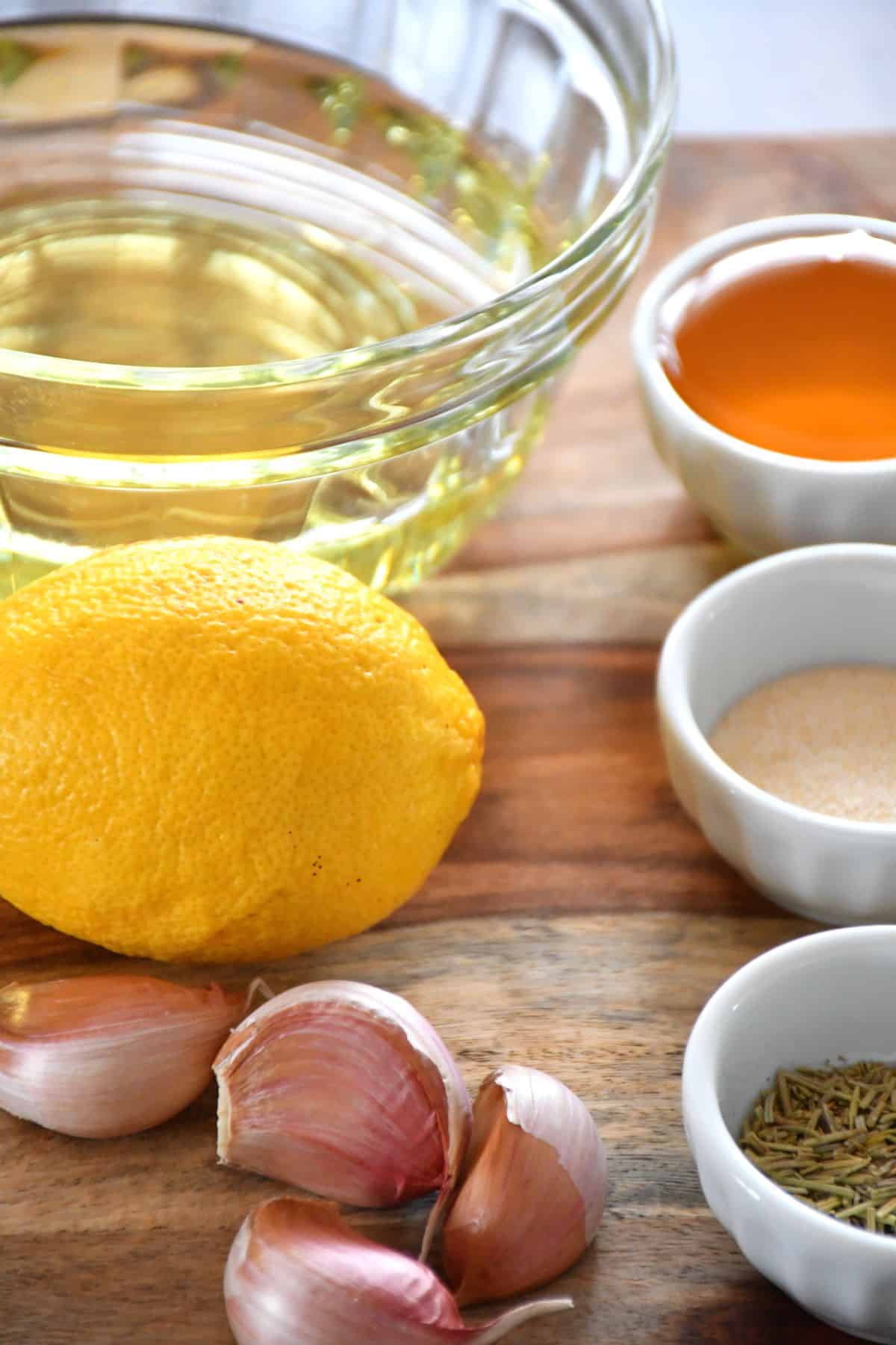 Oil, honey, lemon, garlic, onion powder and dried rosemary displayed on a wooden board to make honey and rosemary marinade.