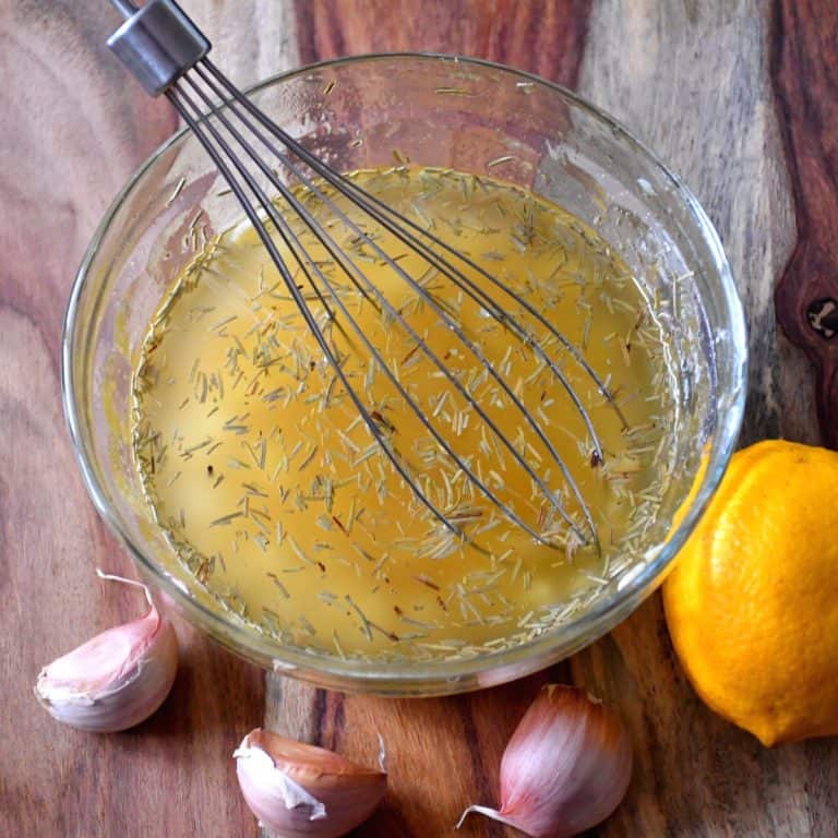 Honey and rosemary marinade in a glass mixing bowl placed on a wooden board with garlic cloves and a lemon.