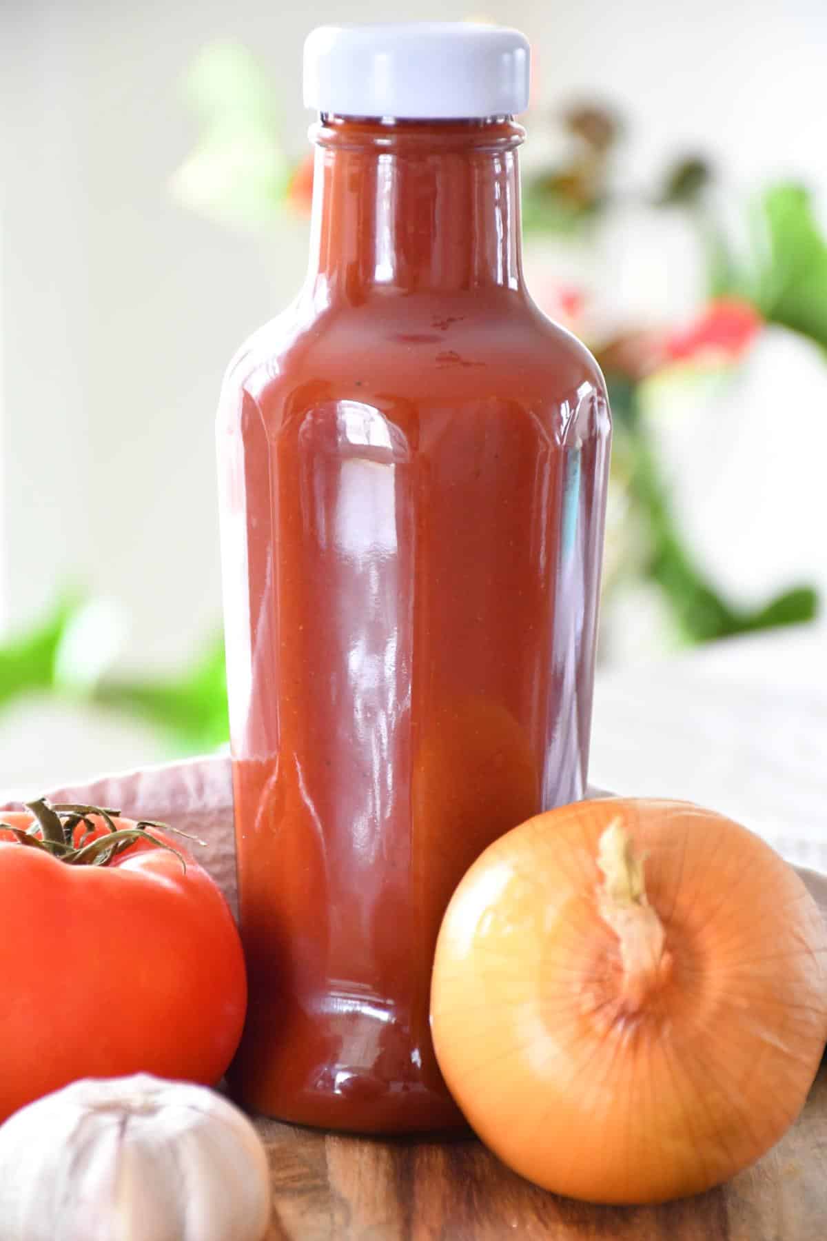Ketchup sans sel ajouté dans une bouteille en verre présentée avec une tomate fraîche, de l'ail et de l'oignon.