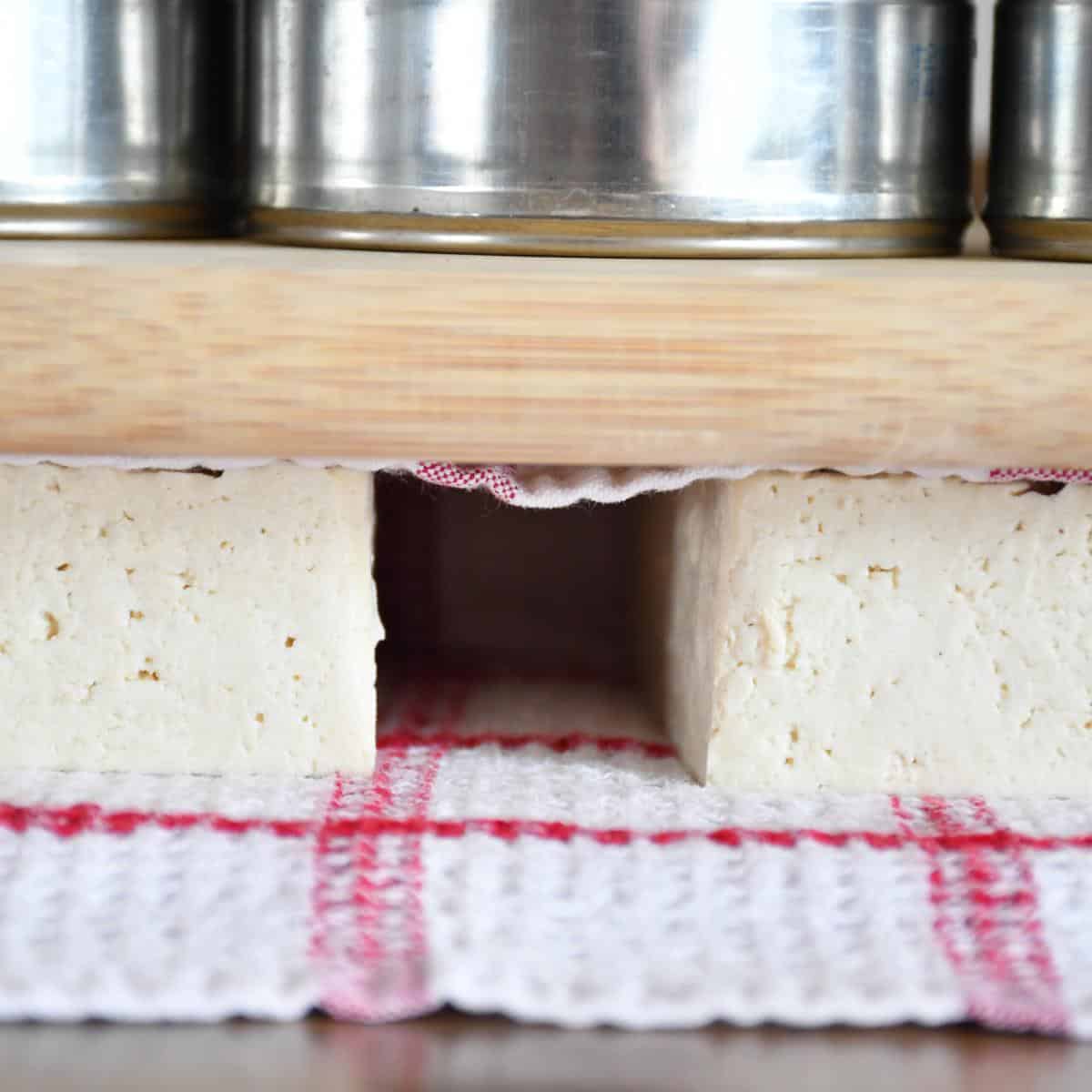 Bloc de tofu pressé entre deux planches à découper pour enlever autant de liquide que possible.
