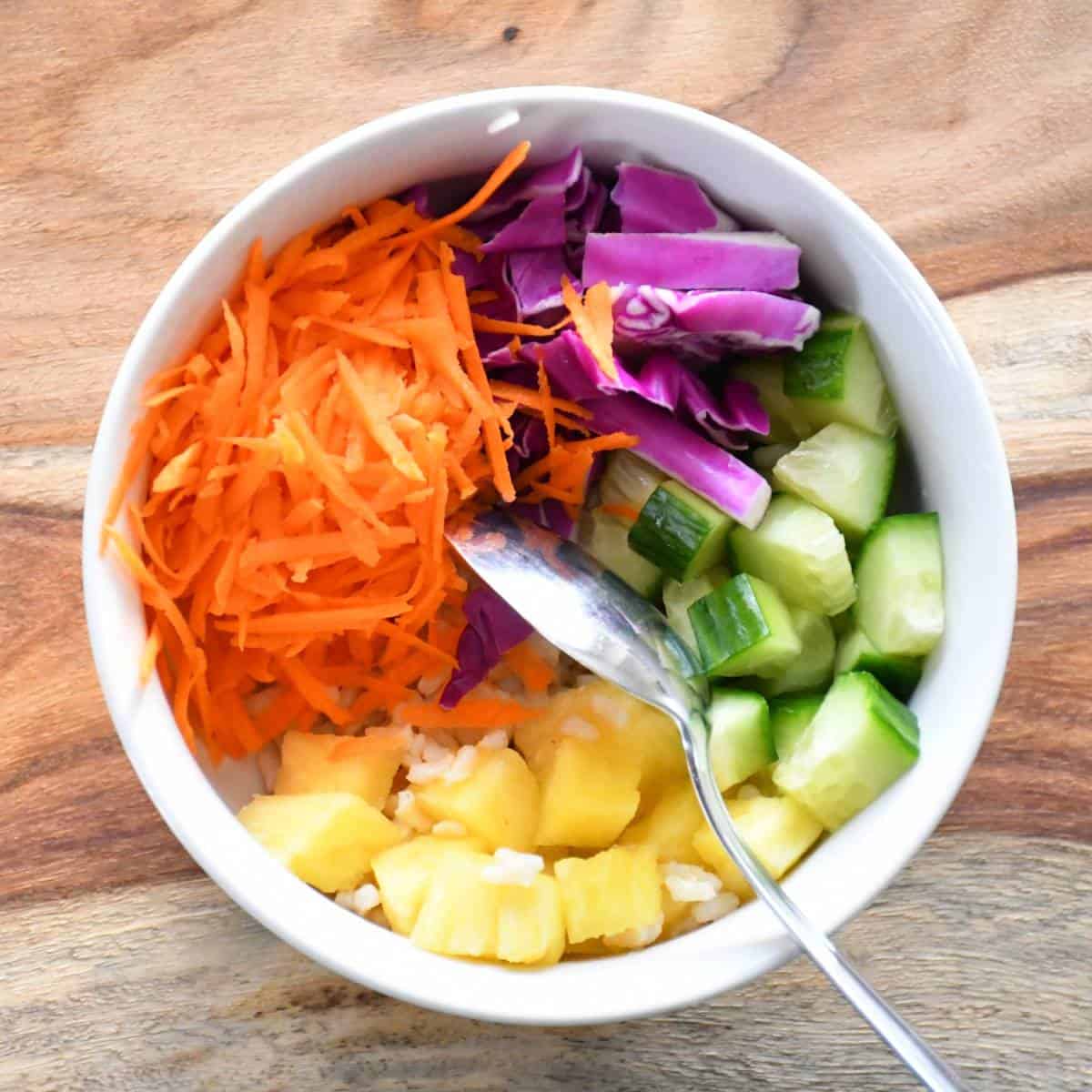 Assembling a colorful vegetarian poke bowl made of brown rice, cucumbers, pineapple, carrots and red cabbage.